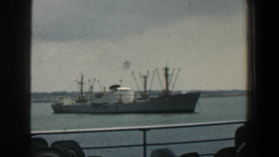 Large cargo vessel with cranes and a white superstructure in Baltimore harbor, viewed past railings in 1961 home-movie footage.
