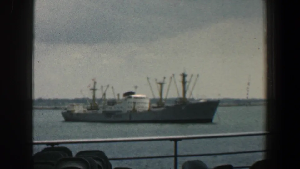1961 Baltimore harbor cargo ship with deck cranes, seen from another vessel with railings and seating silhouettes in the foreground.