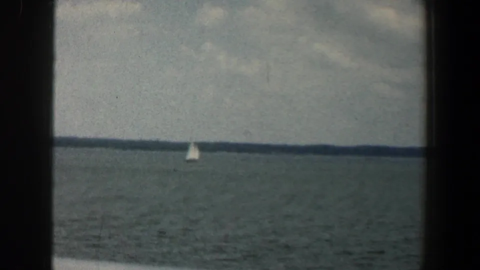 A white sailboat sits near the horizon line on a broad lake, with muted 8mm color and visible film texture.