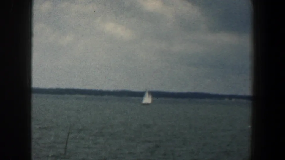 A small white sailboat is framed low on the water beneath a cloudy sky, with a distant shoreline across the background.