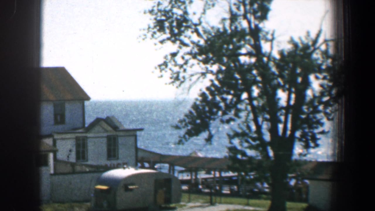 White waterfront house, silver trailer, large tree, and bright water seen through a dark window frame.