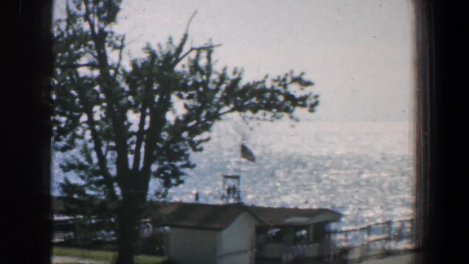 1961 Baltimore-area waterfront frame showing a tree, flag, low building roof, covered picnic area, railing, and reflective water.