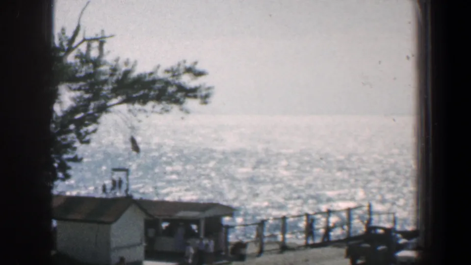 1961 Baltimore-area 8mm frame with a shoreline railing, small waterfront building, flag, bright water, and small indistinct people below.