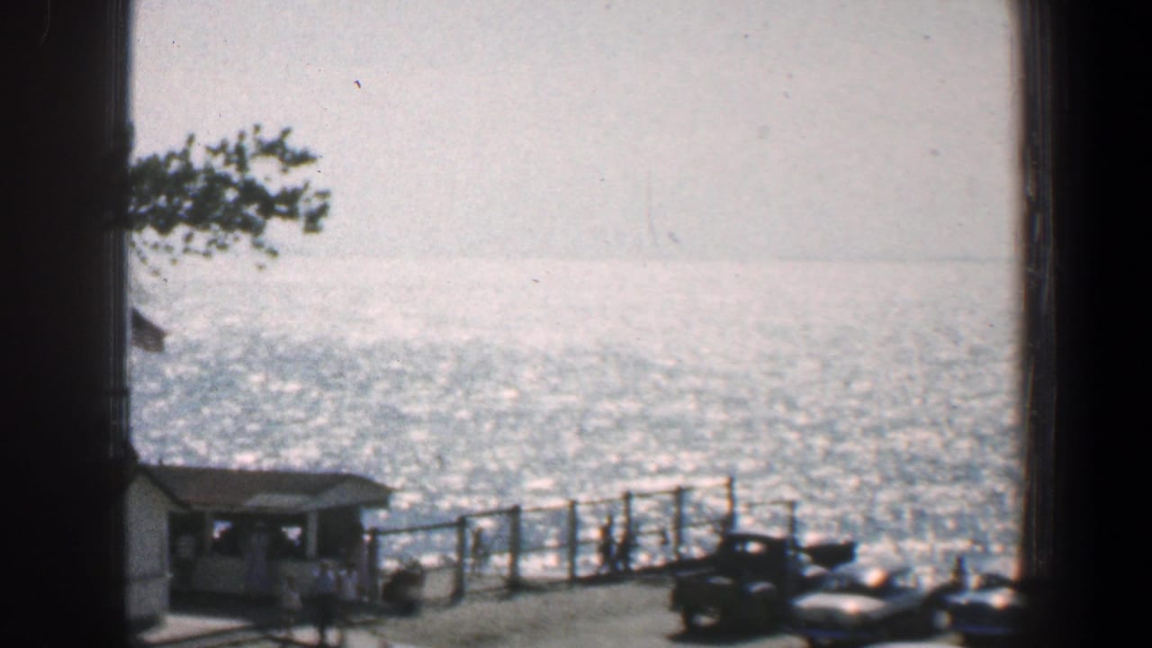 1961 Baltimore home-movie view through a dark window edge toward bright water, pier railing, parked cars, and a small waterfront structure.