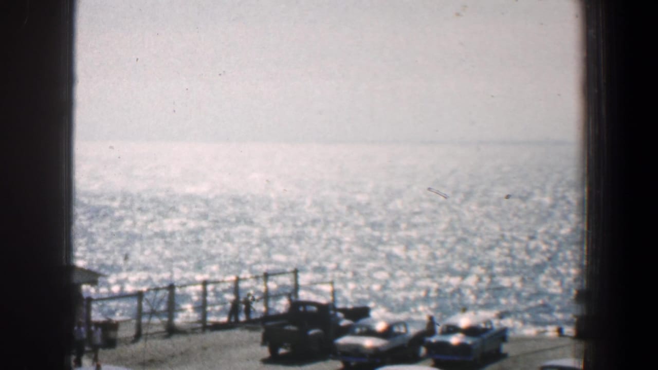 Bright water and pier railing with several parked cars seen through dark vertical window framing.
