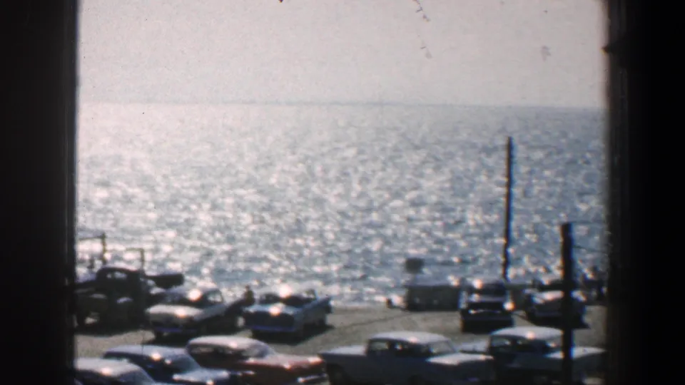 1961 Baltimore waterfront parking area with classic cars, vertical posts, bright water, and dark interior framing from the camera position.