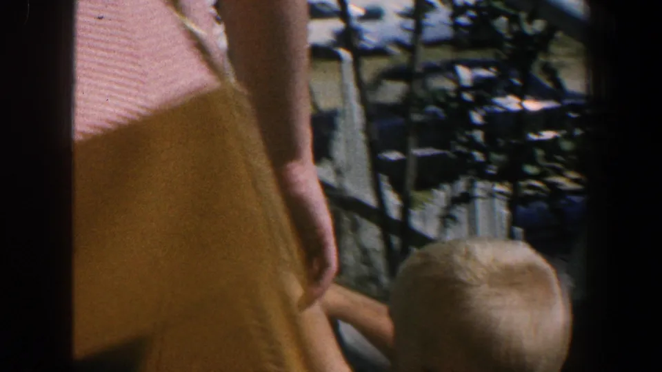 1961 Baltimore home-movie frame showing a partial adult and child near the camera, with parked cars and waterfront surroundings beyond them.