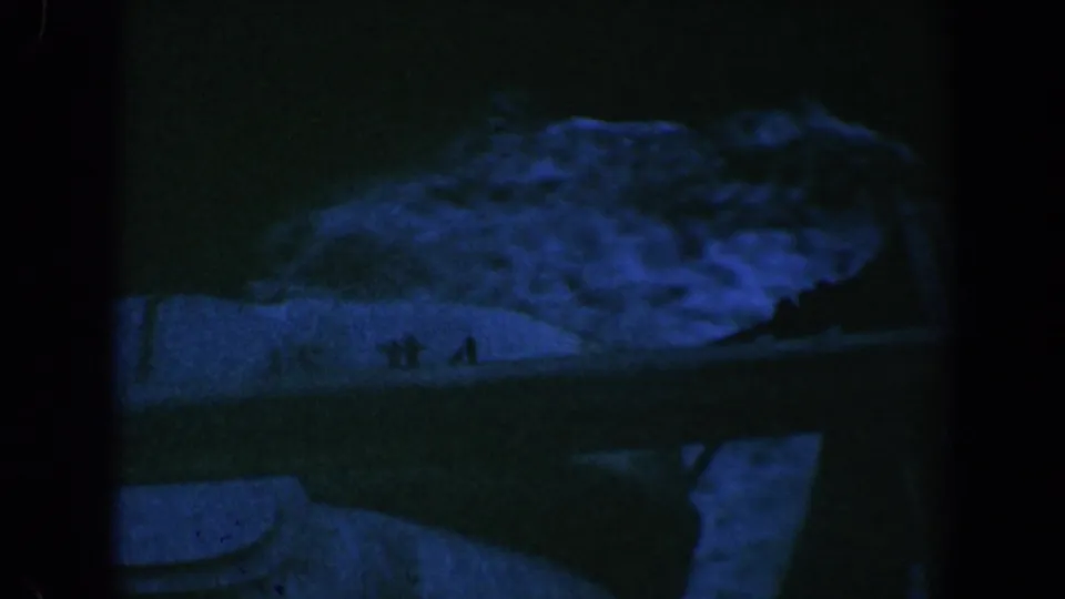 Low-light 1961 Baltimore 8mm frame showing a pale indistinct form on or near dark water, with blue-green cast and heavy grain.