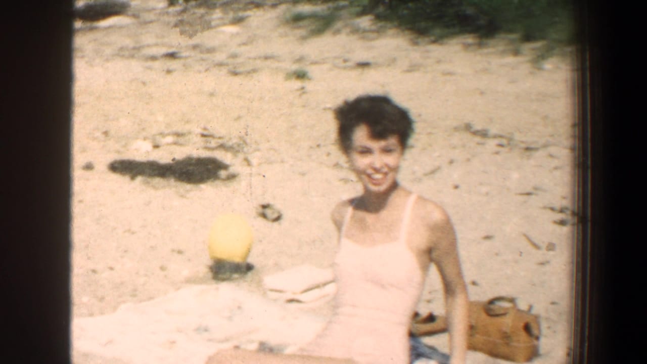 In this 1961 Baltimore, Maryland 8mm home-movie frame, a young woman in a light swimsuit smiles toward the camera while sitting on sand near a towel, yellow beach ball, and tan bag.