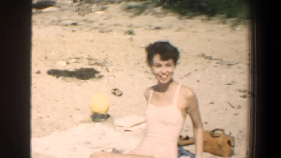In this 1961 Baltimore, Maryland 8mm home-movie frame, a young woman in a light swimsuit smiles toward the camera while sitting on sand near a towel, yellow beach ball, and tan bag.