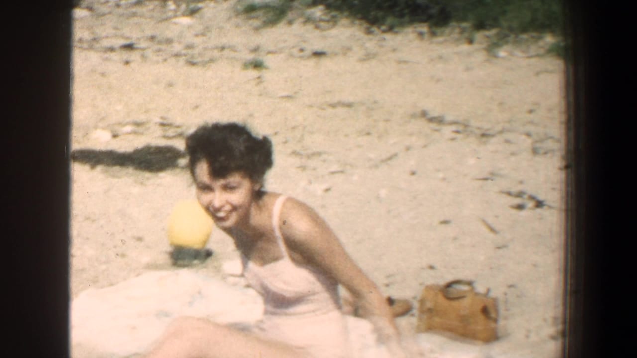 Young woman leans forward laughing on a sandy beach with a yellow ball and bag nearby.