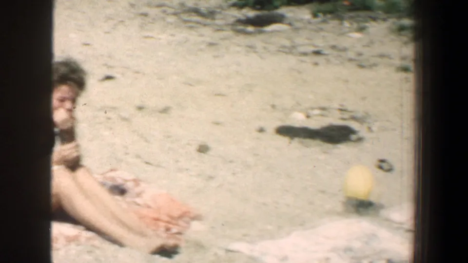 A partly cropped 8mm frame shows a woman reclining on a towel at the edge of the shot, with sandy ground and a yellow beach ball occupying the center-right of the beach scene.