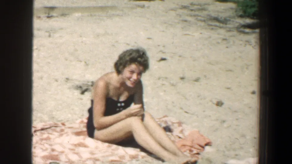 In the final 1961 beach frame, a woman in a dark swimsuit sits on a patterned towel on the sand and smiles toward the camera, with scattered stones and beach debris around her.
