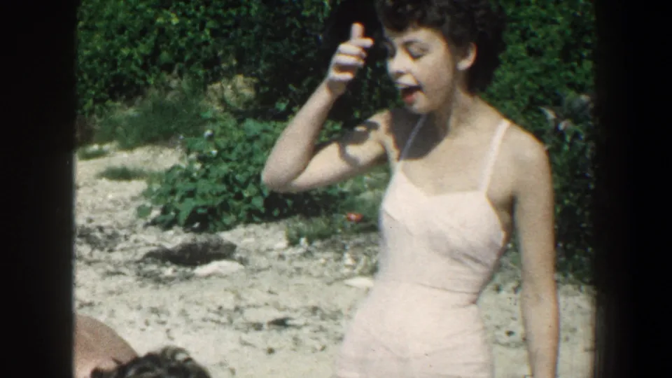 In 1961 Baltimore home-movie footage, a woman in a pink swimsuit adjusts her dark hair on a bright beach.