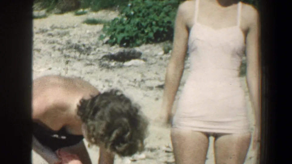 A person with curly hair bends over an orange towel while a woman in a pink swimsuit stands nearby on the beach.