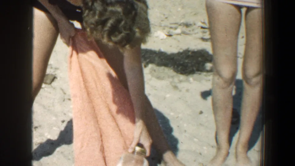 A person bends over an orange towel on the sandy beach while the standing woman in the pink swimsuit is partly framed at right.