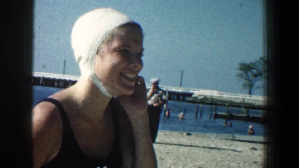 In supplied 1961 Baltimore beach footage, a woman in a white swim cap laughs beside the water with a distant pier behind her.