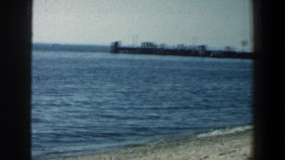 A waterfront view from the supplied Baltimore beach scene shows small waves, sandy shore, and a distant pier in faded 8mm color.