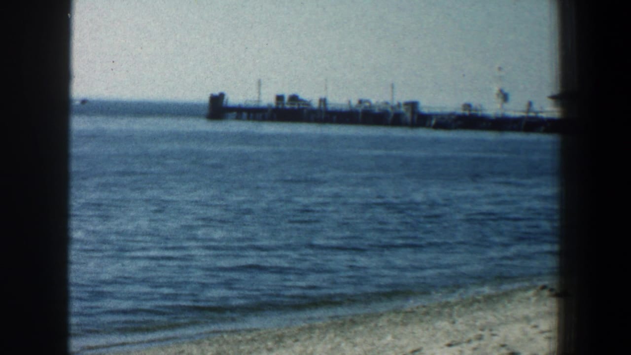 1961 Baltimore beach scene on 8mm film showing sandy shore, blue water, and a long pier across the background.