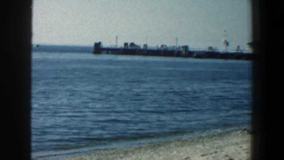 1961 Baltimore beach scene on 8mm film showing sandy shore, blue water, and a long pier across the background.