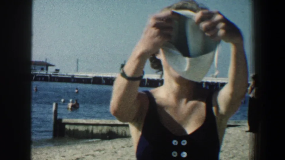A woman in a dark one-piece swimsuit raises a swim cap toward the camera, with beach, swimmers, and pier behind her.