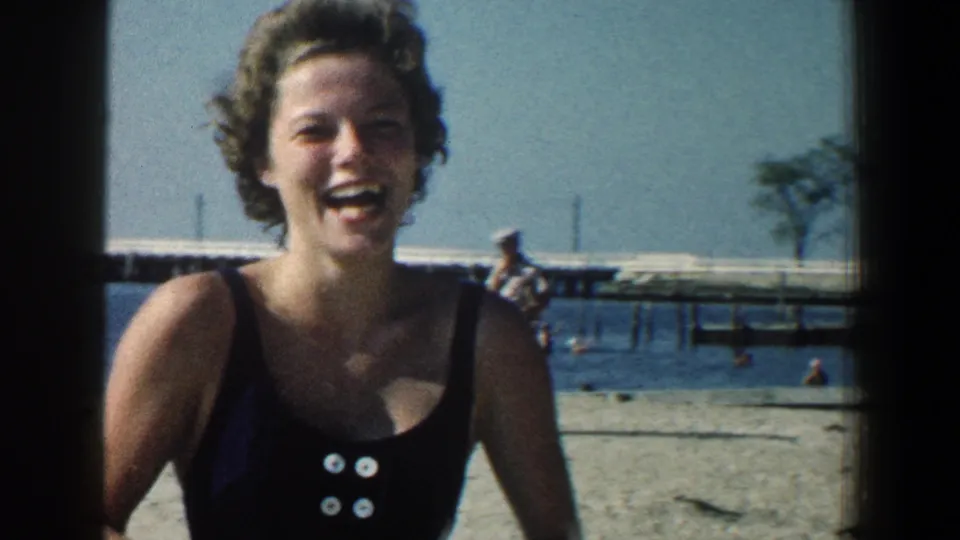 A smiling woman in a dark swimsuit looks toward the camera on a Baltimore beach, with swimmers and a pier in the background.