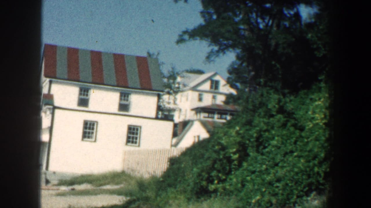 1961 8mm frame of a white Baltimore-area seaside hotel exterior with a striped roof, trees, and sandy foreground.