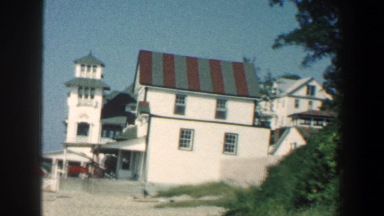 White hotel exterior with tower, striped roof, porch area, and greenery.