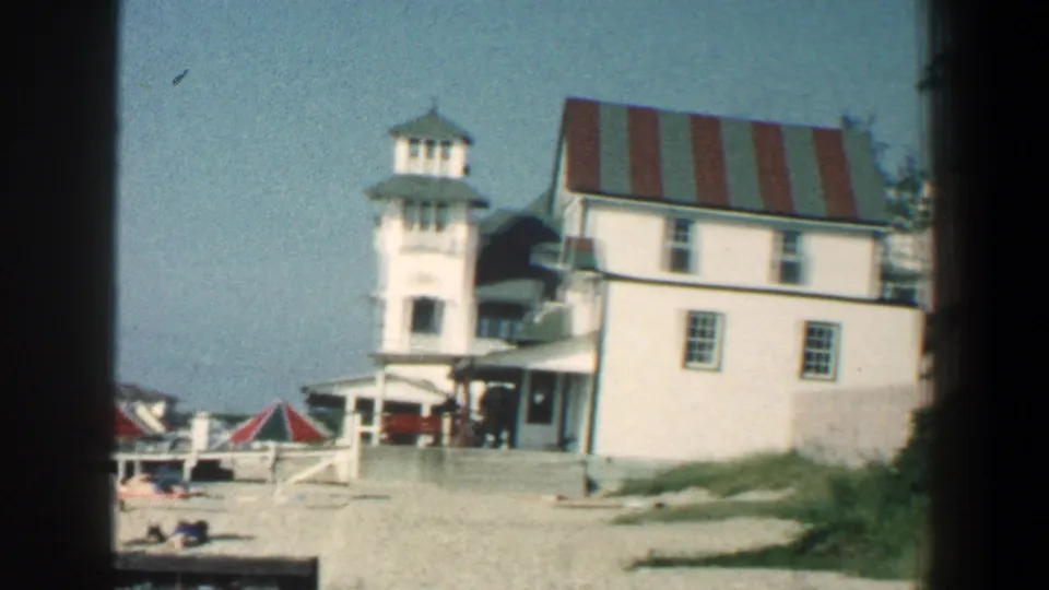 1961 8mm frame of the Baltimore-area hotel and beach, with red-and-white umbrellas and distant beachgoers on the sand.