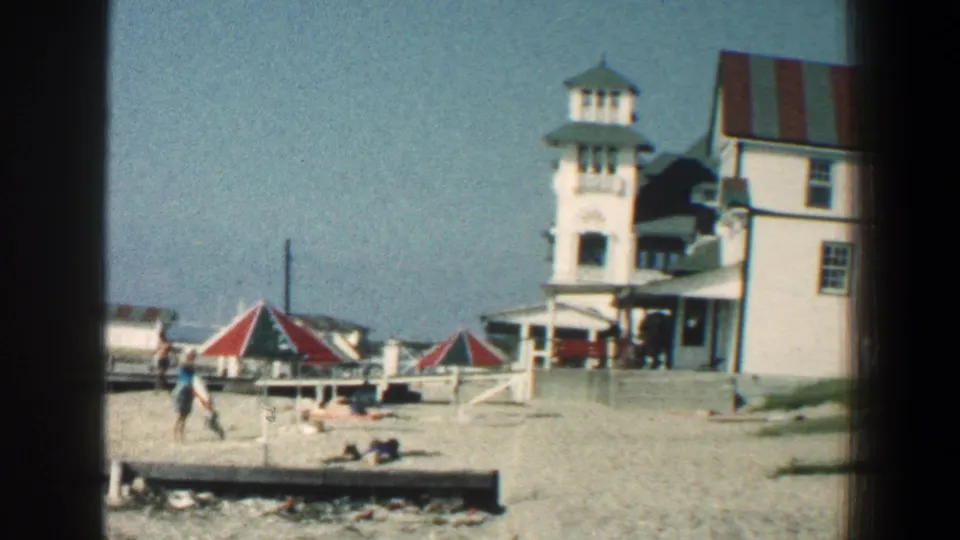 1961 8mm frame showing the Baltimore-area seaside hotel, beach umbrellas, sandy foreground, and distant people near the shore.