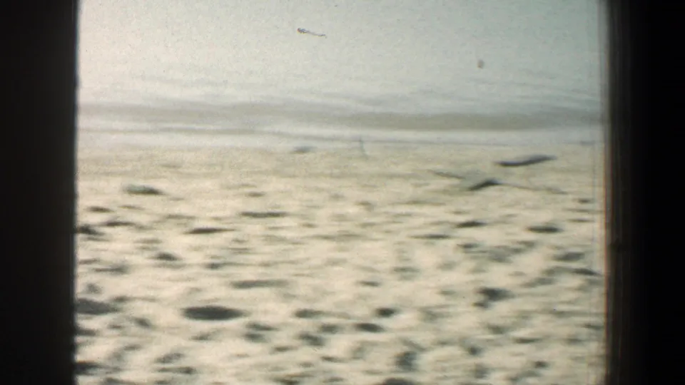 Bright 1961 8mm beach frame showing pale sand, shallow water, and a hazy shoreline horizon.