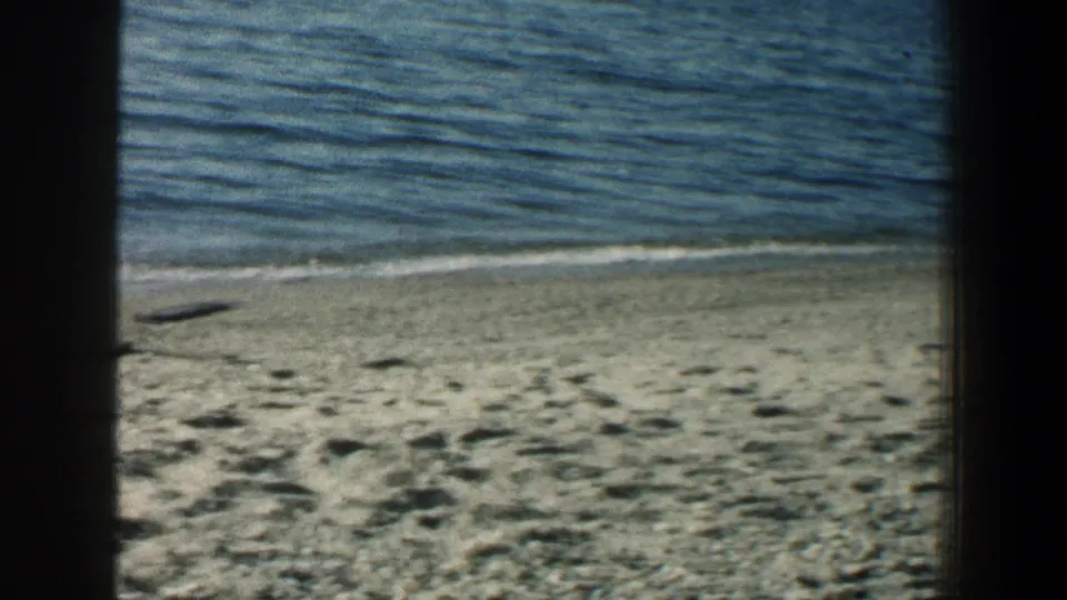 1961 Baltimore beach view with textured sand, gentle water, and a distant pier along the right horizon.