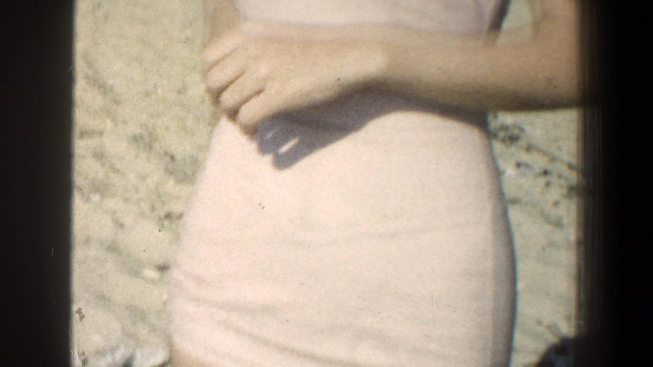 1961 Baltimore-area home-movie frame showing a young woman from torso to thigh in a light swimsuit, standing on sand.