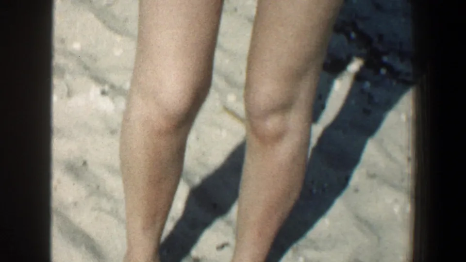 1961 Baltimore-area beach frame showing bare legs and the lower edge of a swimsuit against sunlit sand.