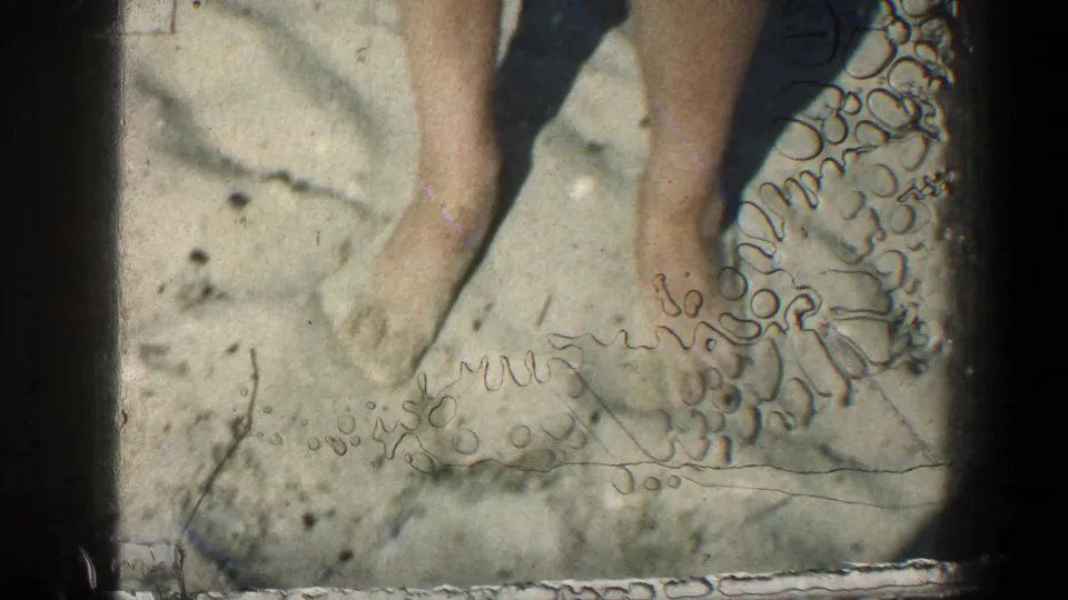 Home-movie beach close-up from 1961 showing bare feet in sand, lower legs, shadows, and visible film wear.