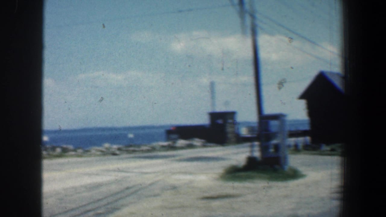 Gravel road leading toward small shoreline buildings and water under a pale sky.