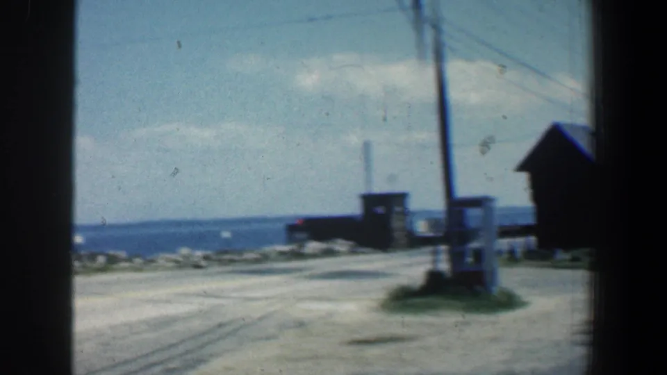 1961 8mm coastal Maryland road scene with small waterfront structures, a utility pole, rocks, and open water beyond.