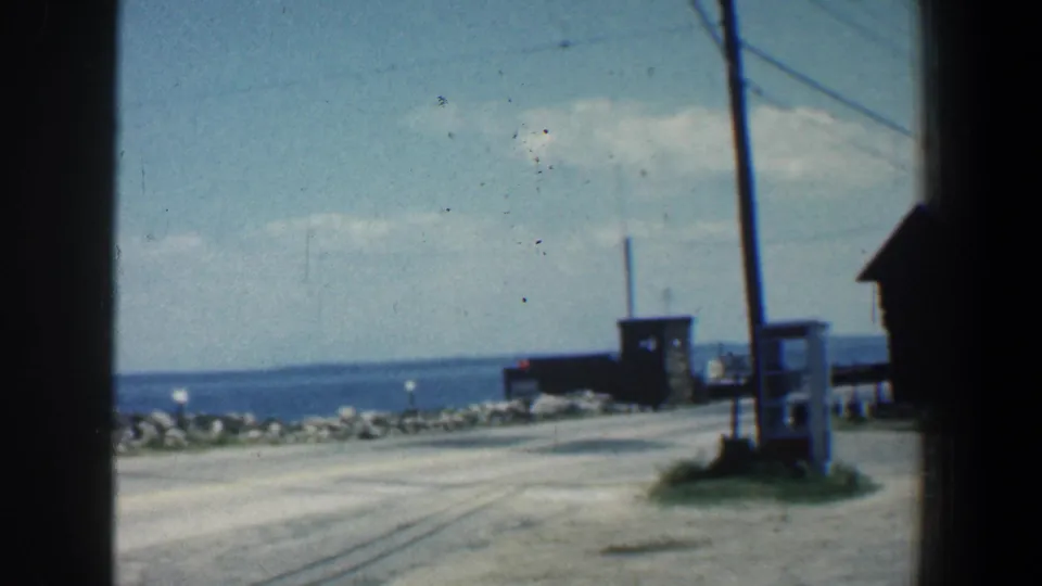 1961 8mm frame showing a coastal road in Maryland with utility poles, small shoreline buildings, rocks, and distant water.
