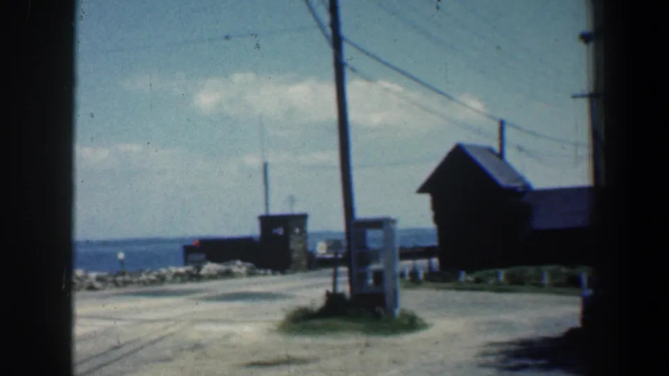 1961 8mm Maryland shoreline view with a gravel road, utility poles, small wooden structure, rocky breakwater, and open water.