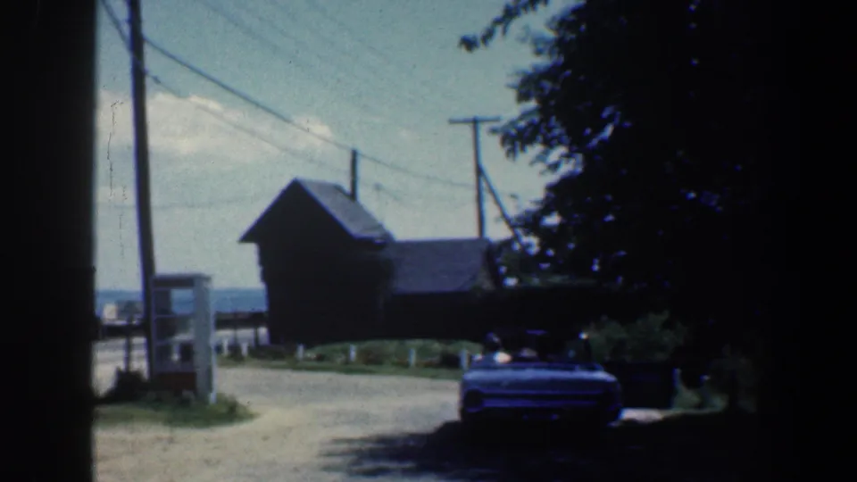 1961 Baltimore-area frame with a classic convertible in tree shade near a roadside phone booth and distant waterfront.