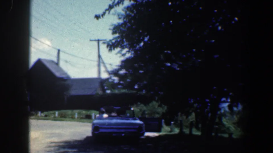 1961 Baltimore-area 8mm frame of a classic convertible under trees beside a road and small wooden building.