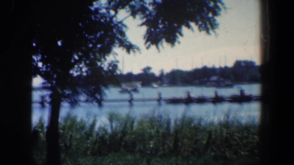 1961 Baltimore-area 8mm frame with small boats near a wooden pier, shoreline grasses in front, and tree branches overhead.