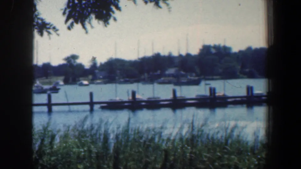 1961 Baltimore-area waterfront view with moored small boats, a wooden pier line, reeds, tree shade, and distant shoreline structures.