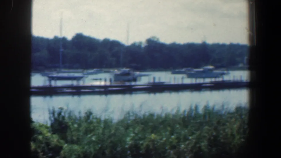 1961 Baltimore-area 8mm frame showing a quiet waterfront dock, small boats, reeds, and a wooded far shore.