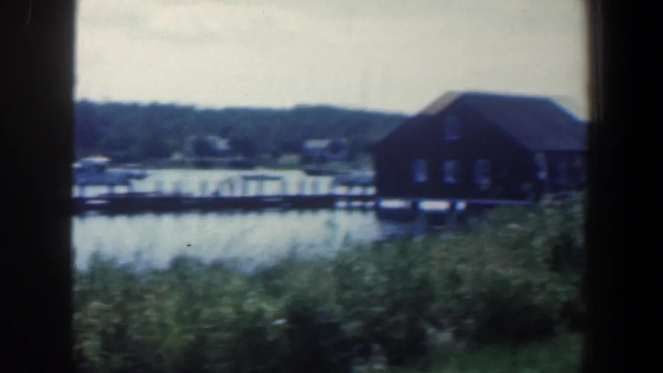 1961 Baltimore-area frame with a dark house on pilings near a dock, small boats, calm water, and distant trees.