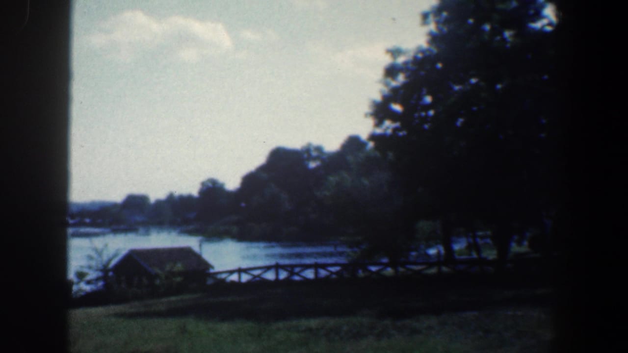 Calm lake with a small shoreline structure, rustic fence, trees, grass, and dark frame edges.