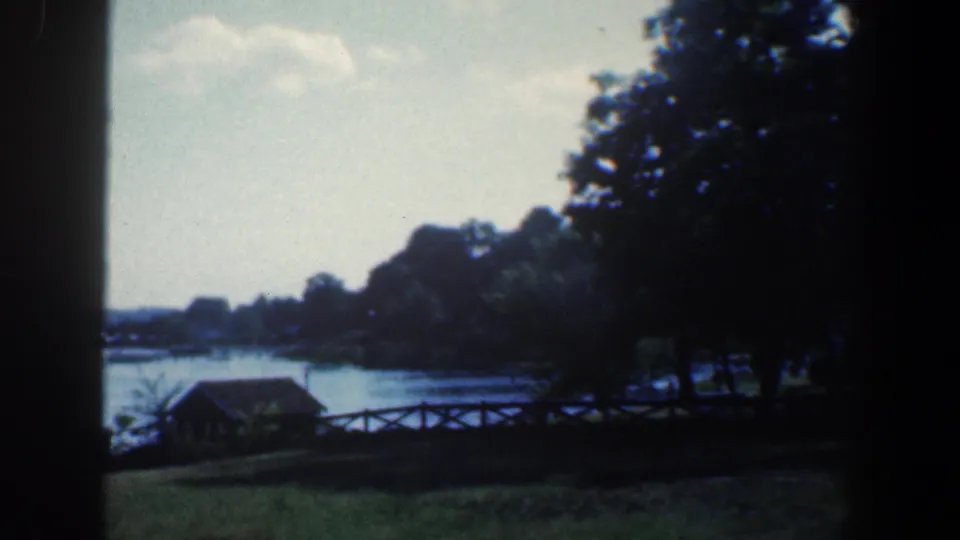 1961 8mm Baltimore-area lakeside view with a small wooden structure, fence, grassy bank, trees, and calm water seen through dark edges.