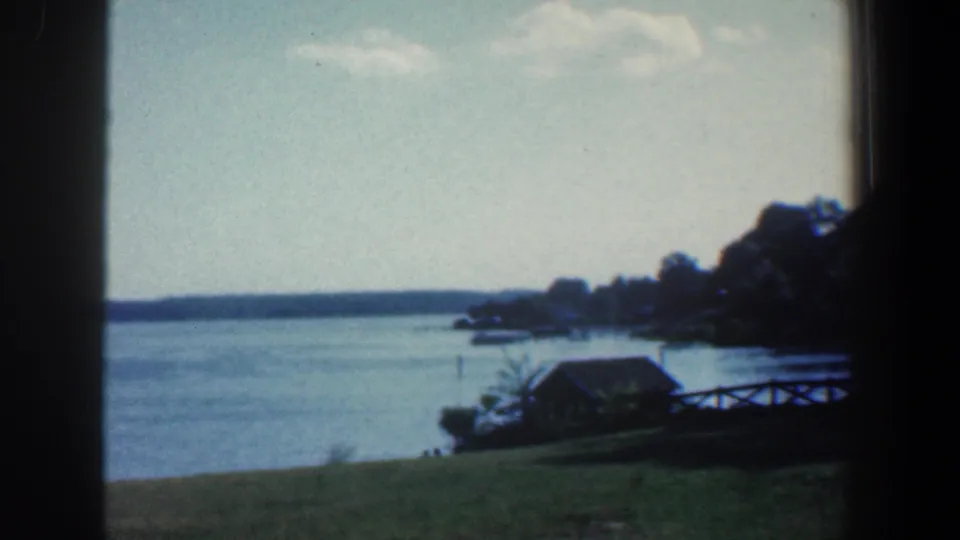 1961 8mm lakeside panorama near Baltimore with open water, a grassy bank, small wooden structure, fence, and tree-lined distant shore.