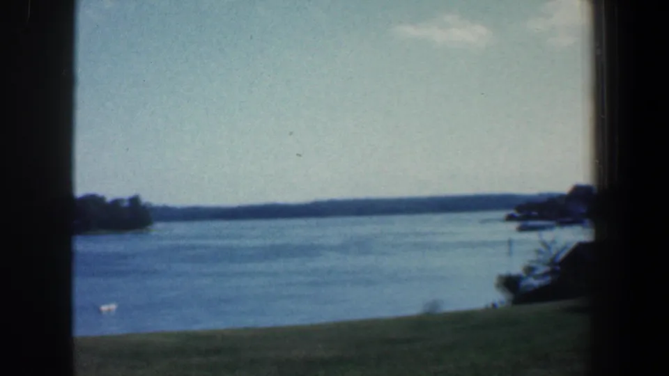 1961 Baltimore-area 8mm frame with a broad lake surface, low grassy shore, distant horizon, tree mass at right, and dark film edge.