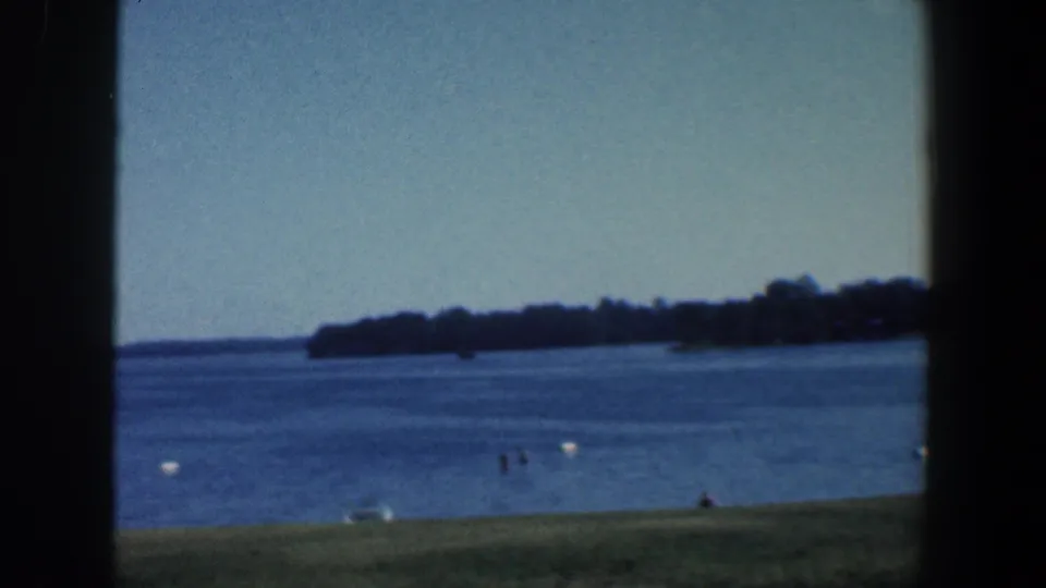 1961 Baltimore-area 8mm view across a lake, with a distant wooded shore, small shoreline figures, buoys, and grass below.